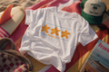 White Philippians 4:4 graphic tee featuring three orange starfish designs photographed on a beach towel with warm coastal sunset lighting.