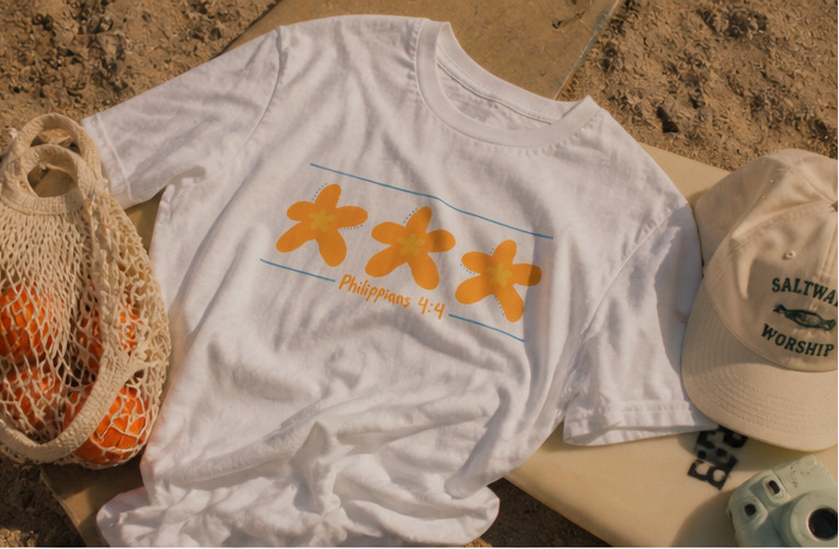 White Philippians 4:4 graphic tee featuring three orange starfish designs photographed on a beach towel in bright coastal sunlight with relaxed summer styling.