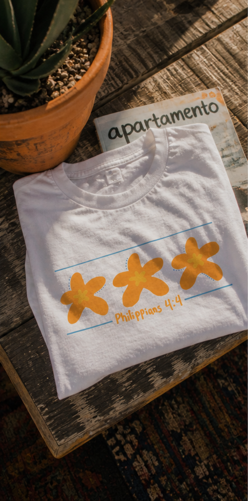 White Philippians 4:4 graphic tee featuring three orange starfish designs photographed on a rustic outdoor table in warm vintage coastal lighting.