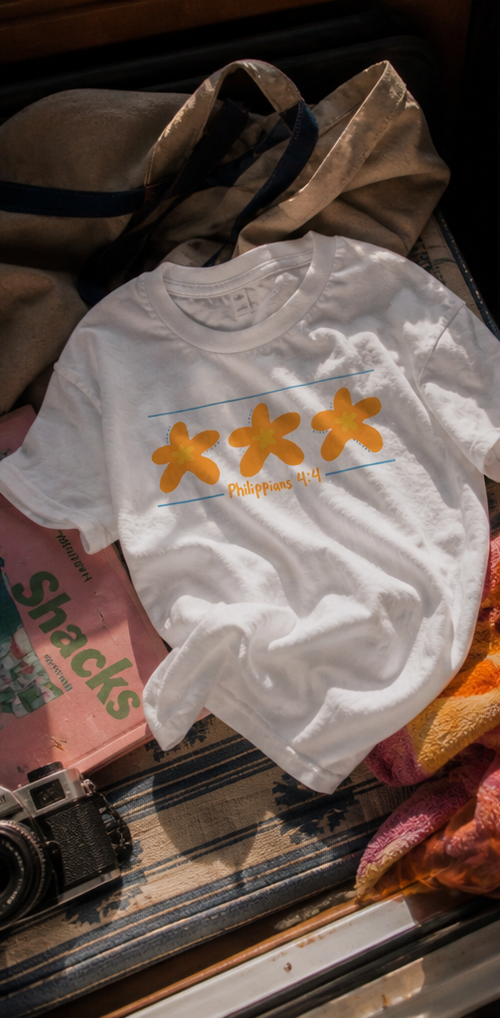 White Philippians 4:4 graphic tee featuring three orange starfish designs photographed in warm coastal sunset lighting with relaxed beach-inspired styling.