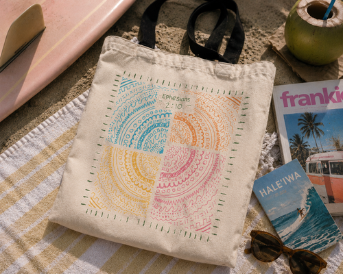 Faith-inspired Ephesians 2:10 tote bag photographed on a beach towel beside a surfboard and coastal accessories in warm sunset lighting.
